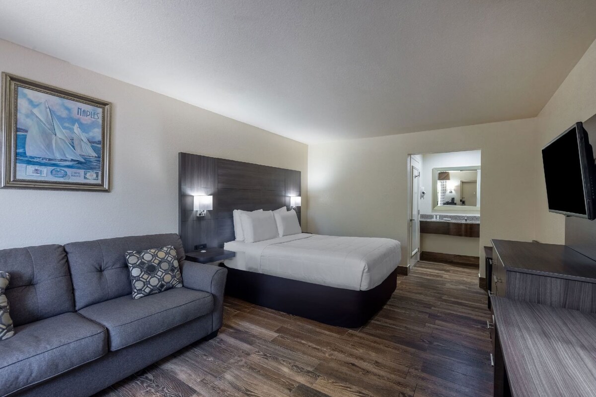 A hotel room with a bed, a gray sofa, a TV, a painting, and a bathroom visible through an open door.