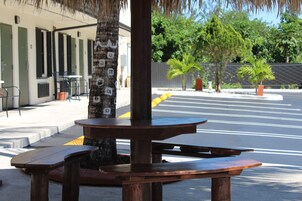 Una mesa de picnic de madera con bancos, un árbol y un edificio con valla al fondo.
