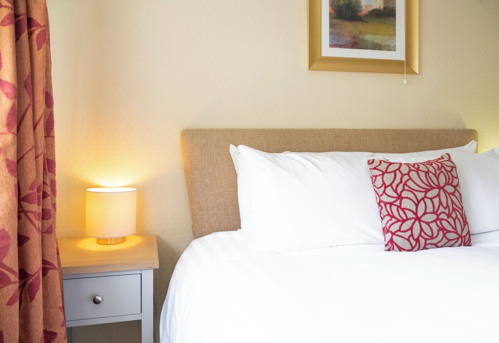 Ein Hotelzimmer mit einem Bett, einem Nachttisch mit Lampe, einem gemusterten Kissen und einem gerahmten Bild an der Wand.