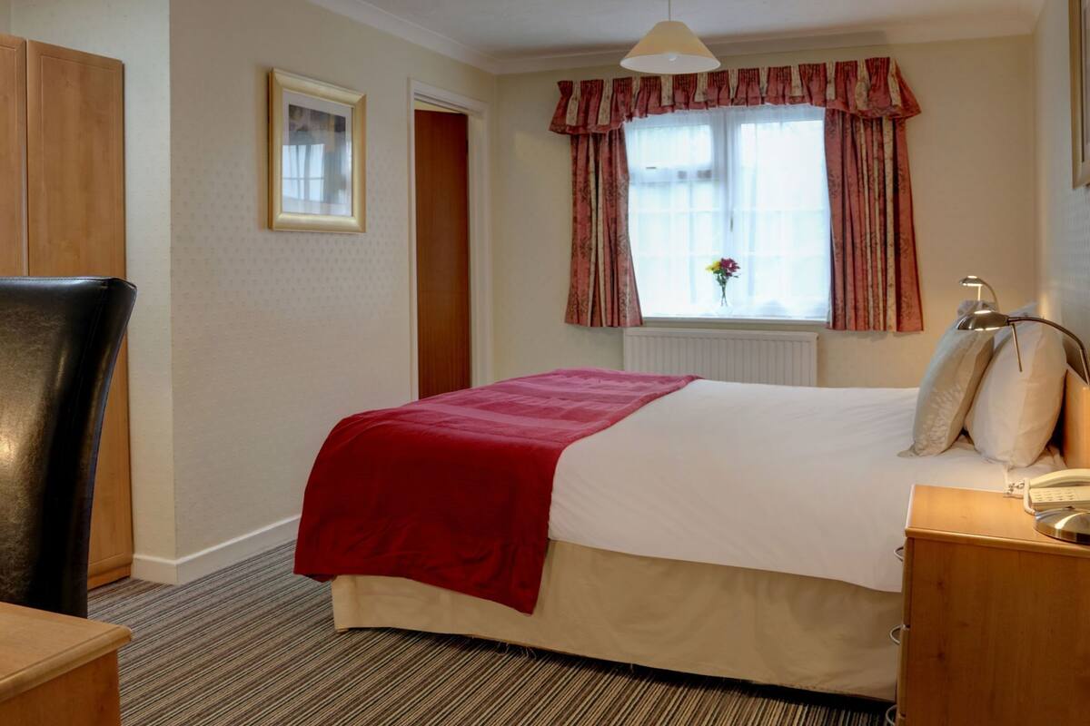 Une chambre d’hôtel comprenant un lit, une chaise, une table de chevet avec une lampe, une fenêtre avec des rideaux et un radiateur.