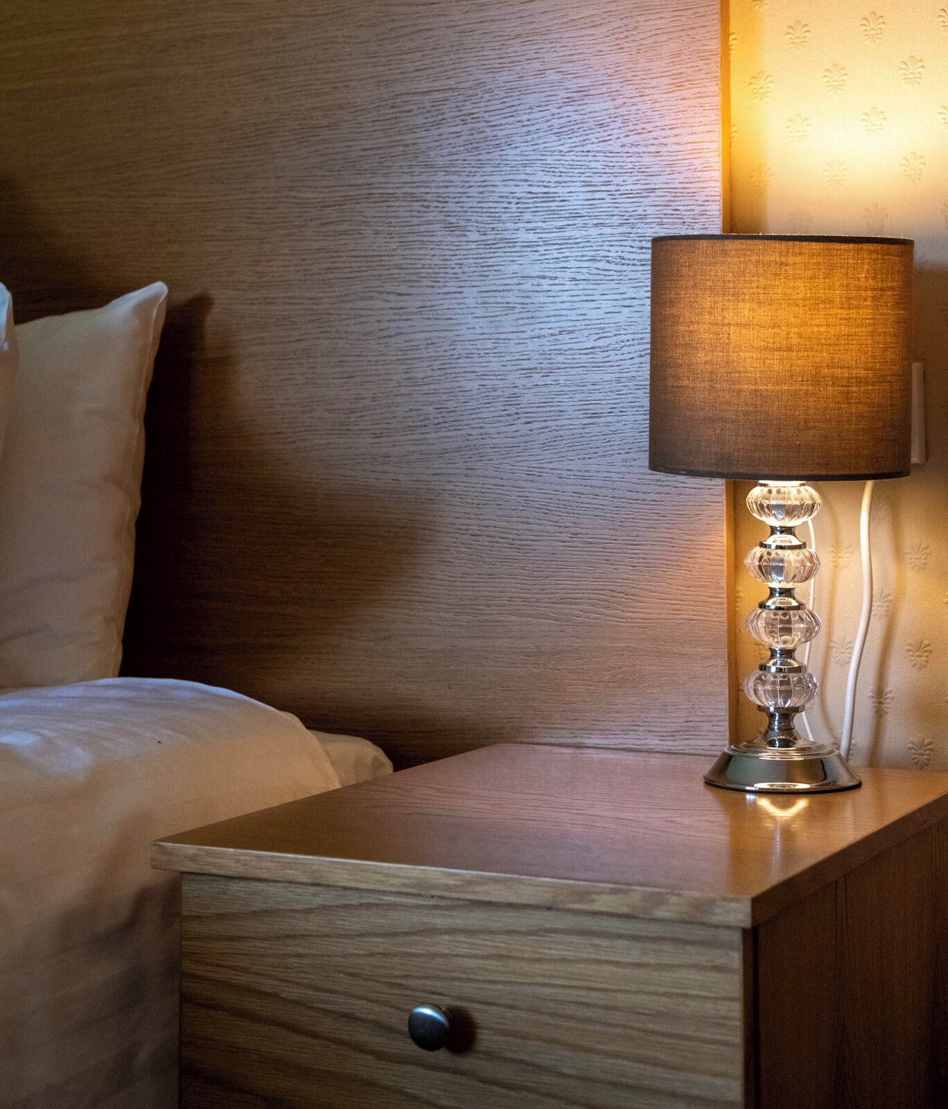 A wooden nightstand with a drawer, a lamp with a textured lampshade, and a bed with white bedding.