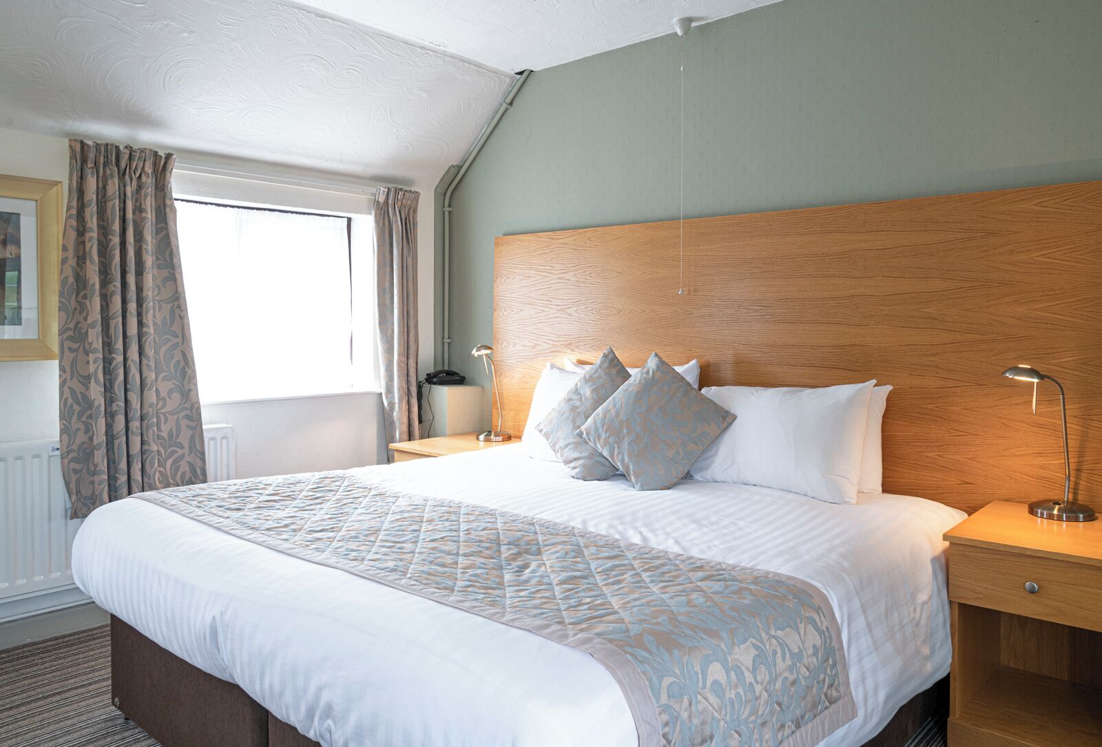 A hotel room with a large bed, two bedside tables, a wooden headboard, and a window with curtains.