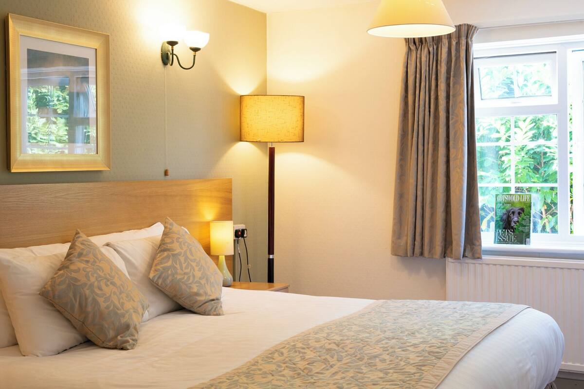 Un lit bien fait, agr émenté d’oreillers à motifs, une lampe de chevet, un tableau encadré au mur et une fenêtre avec des rideaux.
