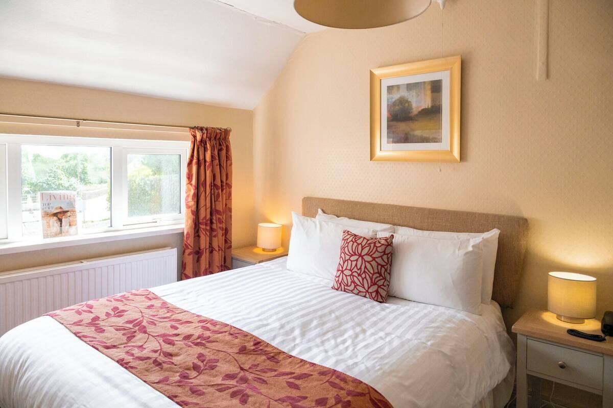 Une chambre à coucher avec un grand lit, une fenêtre avec des rideaux, une table de chevet avec une lampe et un tableau encadré au mur.