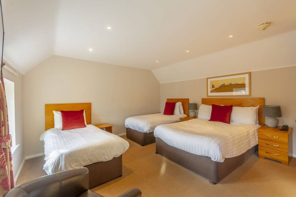 Une chambre d’hôtel avec trois lits, chacun doté d’une tête de lit en bois, de draps blancs et d’oreillers rouges. On y trouve des tables de chevet avec des lampes, une chaise et un tableau encadré au mur.