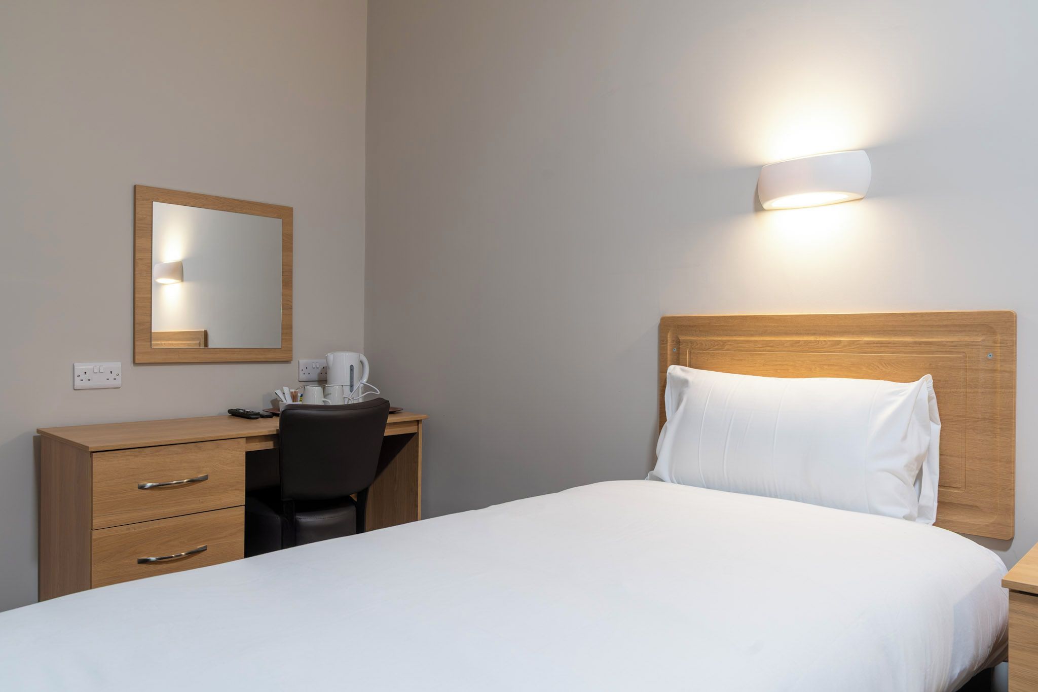 A hotel room with a bed, a desk with a chair, a mirror, and a wall-mounted light.
