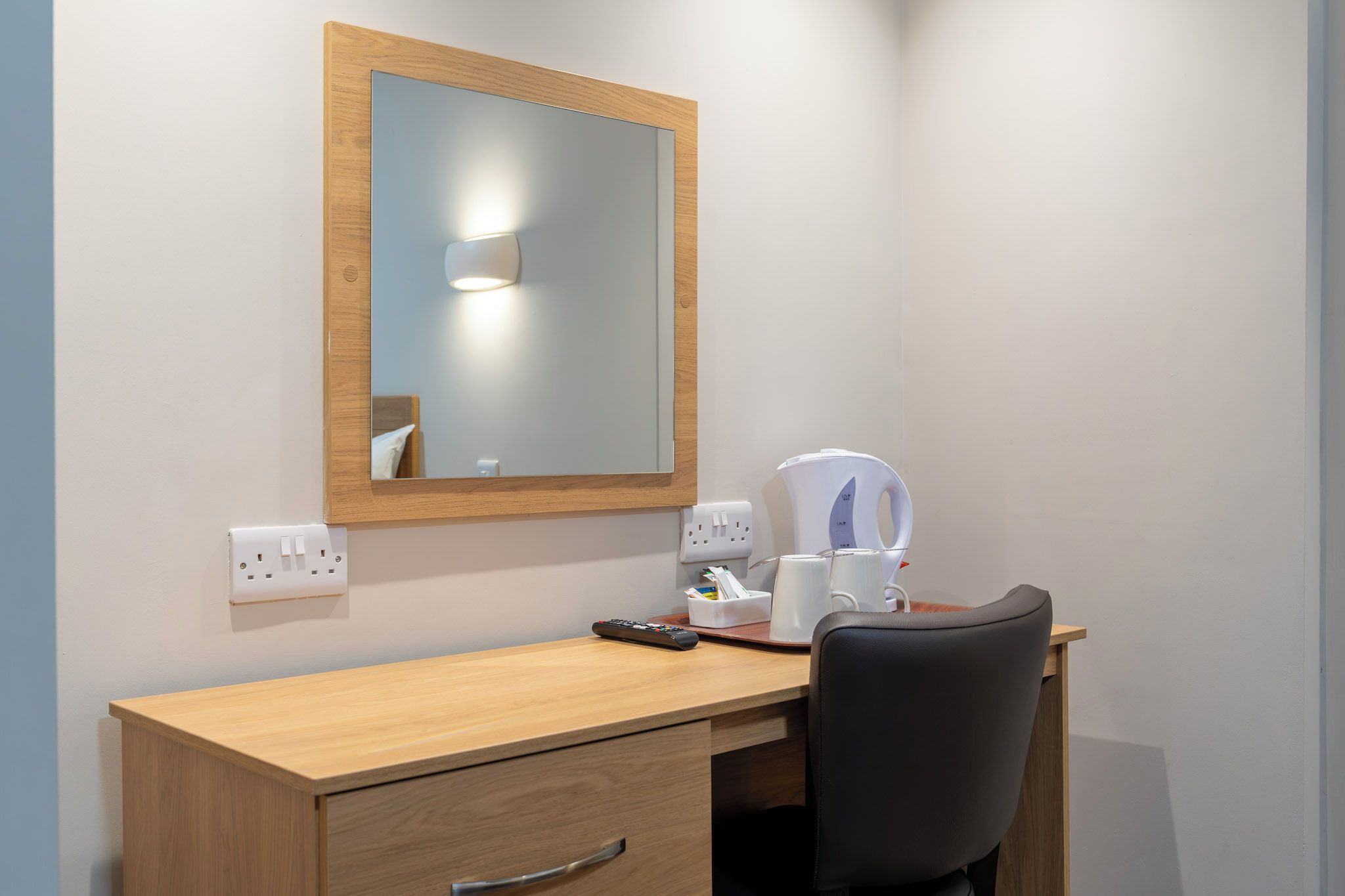 A wooden desk with a mirror, a remote control, a coffee maker, and a chair.