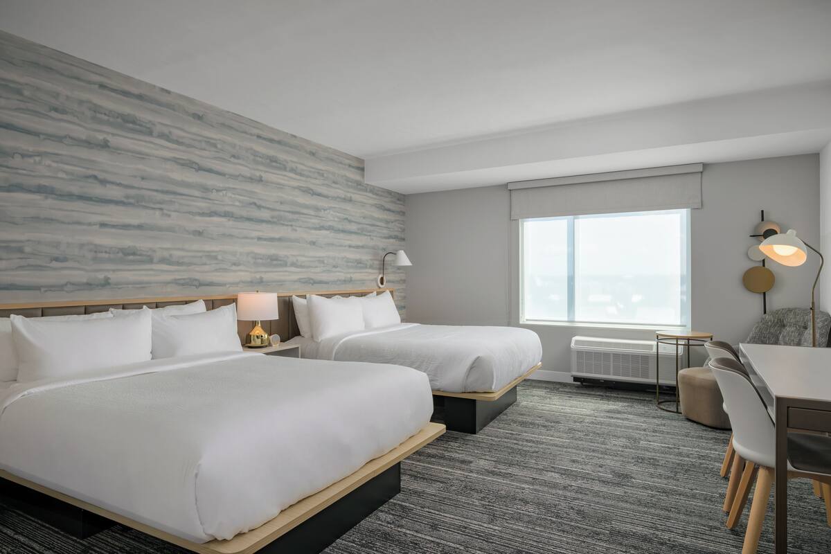 A hotel room with two beds, a desk, a chair, a lamp, and a large window.