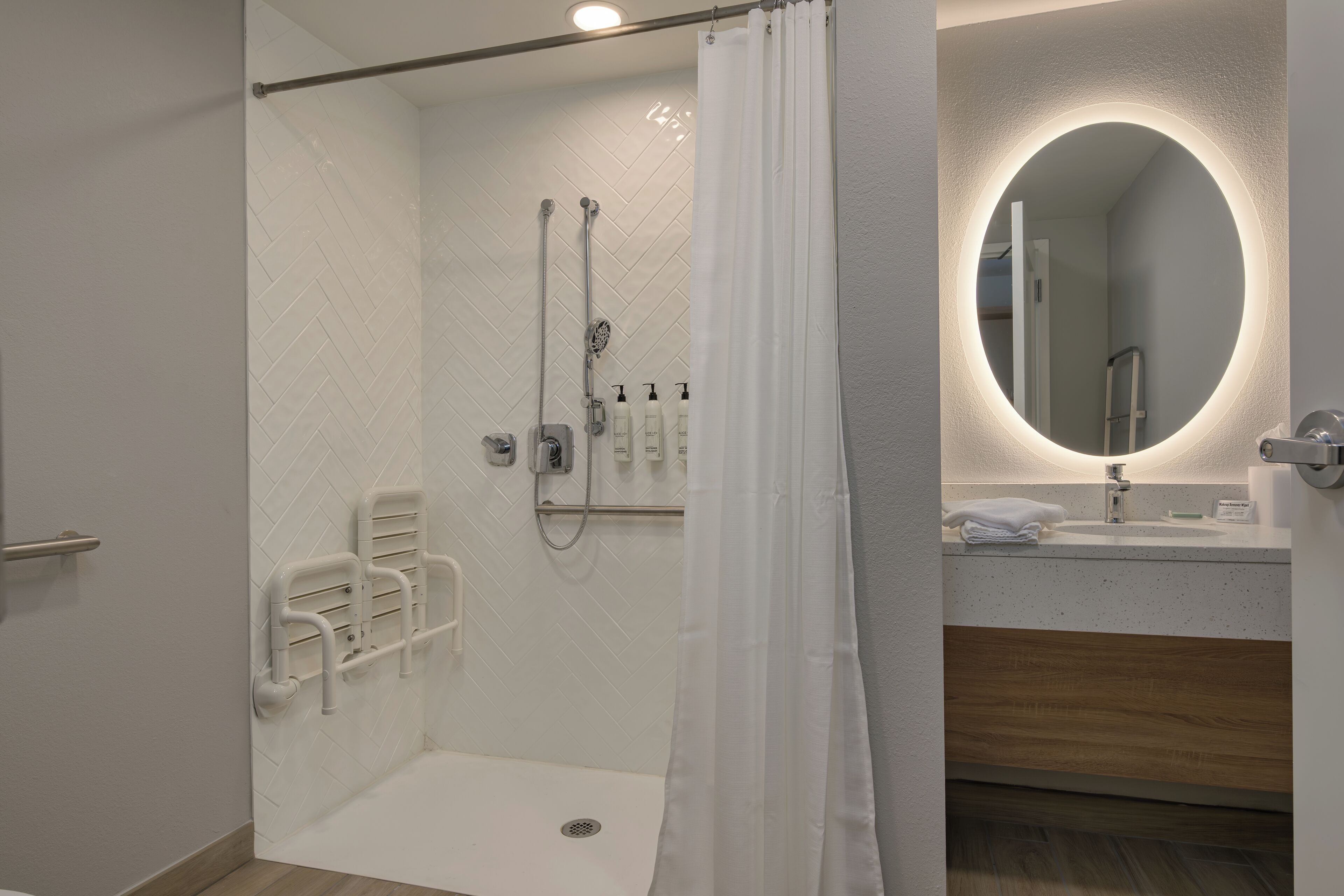 Un baño moderno con ducha envuelta en vidrio, paredes cubiertas de azulejos blancos, un tocador de madera y un espejo redondo con marco iluminado.