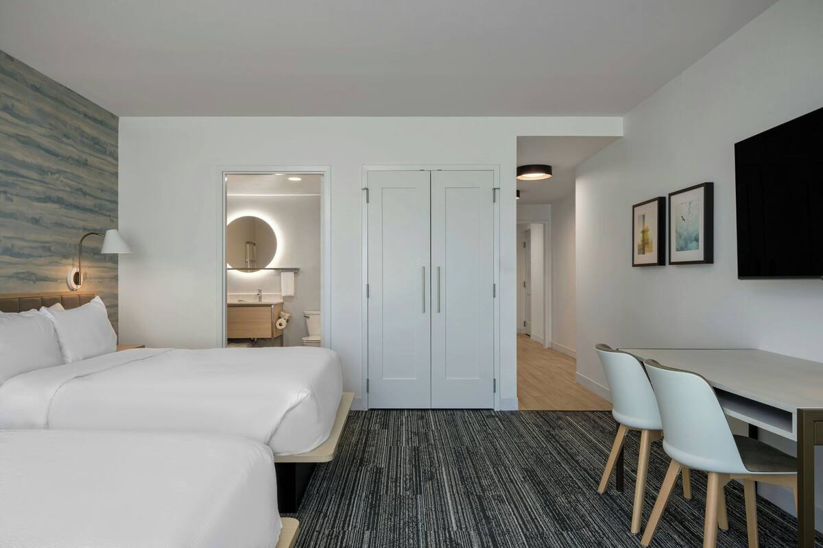 A hotel room with a large bed, a desk with two chairs, a round mirror, and a door leading to a bathroom.