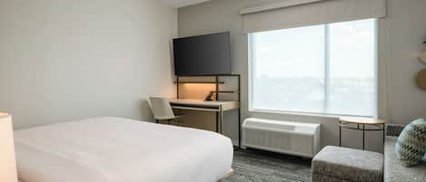 A hotel room with a large bed, a desk with a chair, a television, a window with a view, and a small round table.
