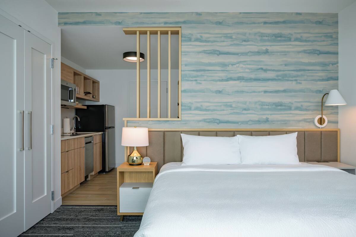 A hotel room with a bed, bedside table, lamp, and a view of the kitchen area.