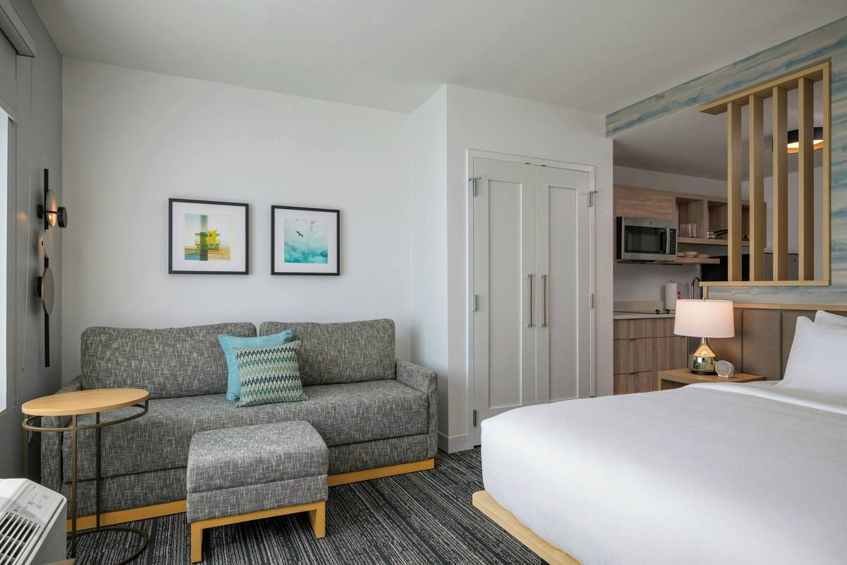 A hotel room with a bed, a sofa, a small table, a microwave, a nightstand with a lamp, and a wardrobe.