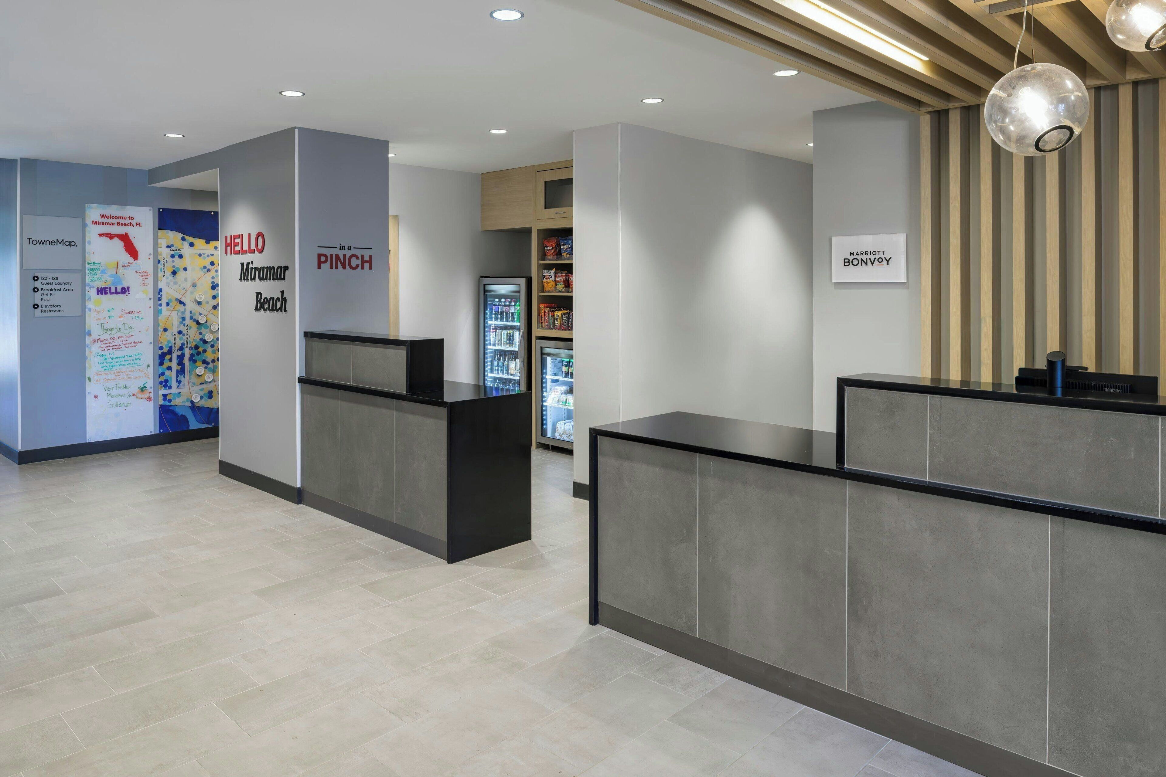 Uma área de recepção moderna de hotel, com balcão de recepção, geladeira abastecida com bebidas e um painel com a inscrição 'Hello Pinch'.
