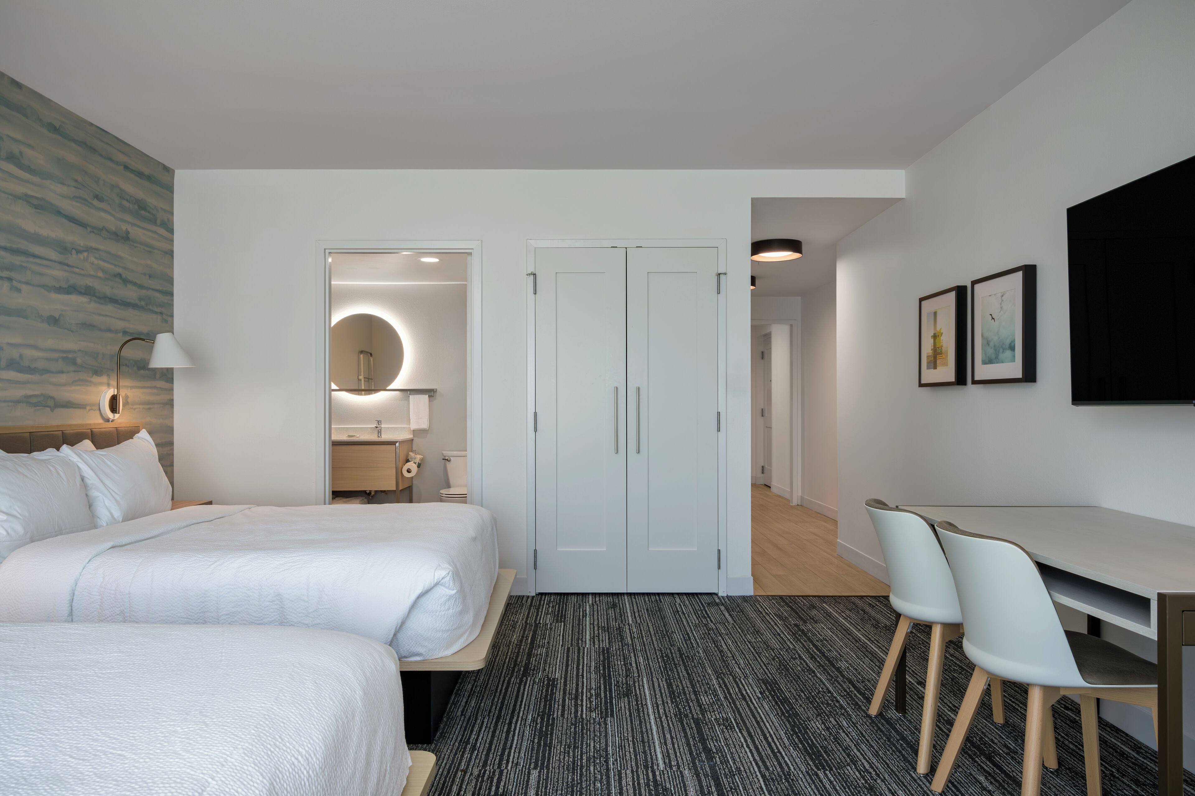 Hotellihuone, jossa on kaksi sänkyä, työpöytä tuoleineen, pyöreä peili ja ovi, joka johtaa kylpyhuoneeseen.
