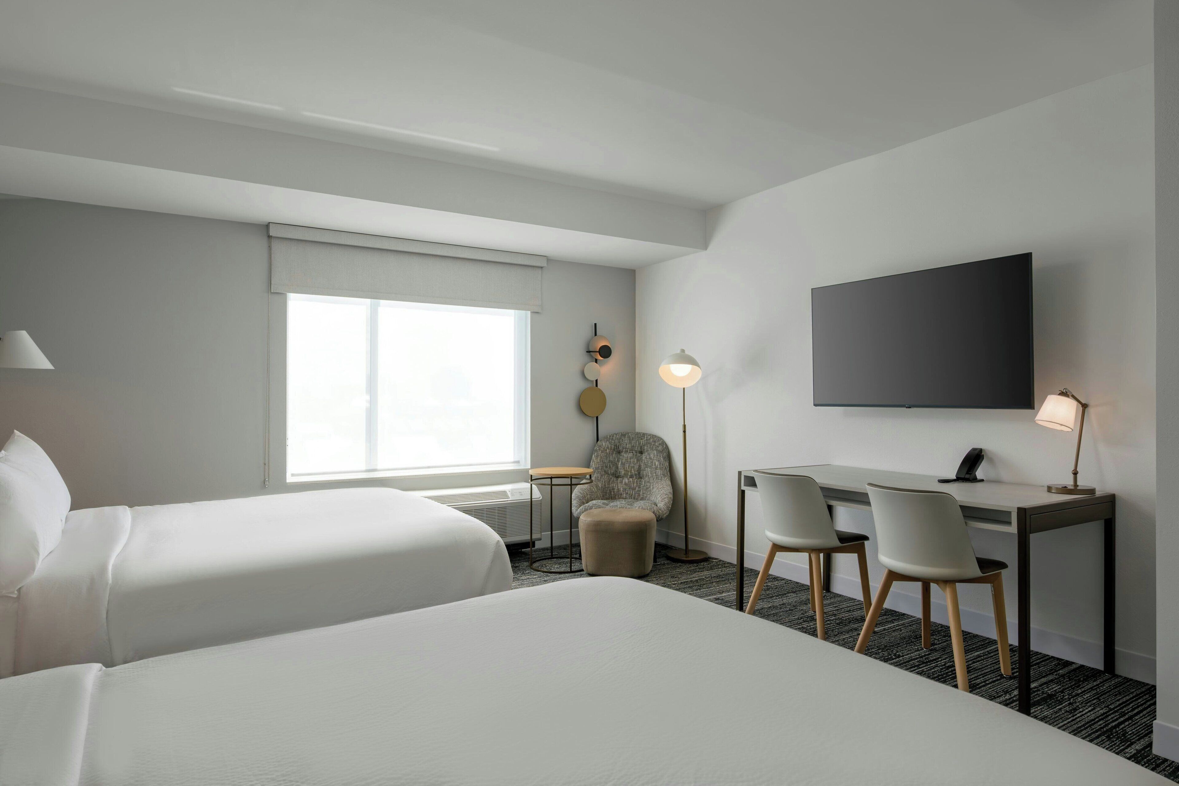 A hotel room with two beds, a desk with chairs, a flat-screen TV, a lamp, and a window with a view.