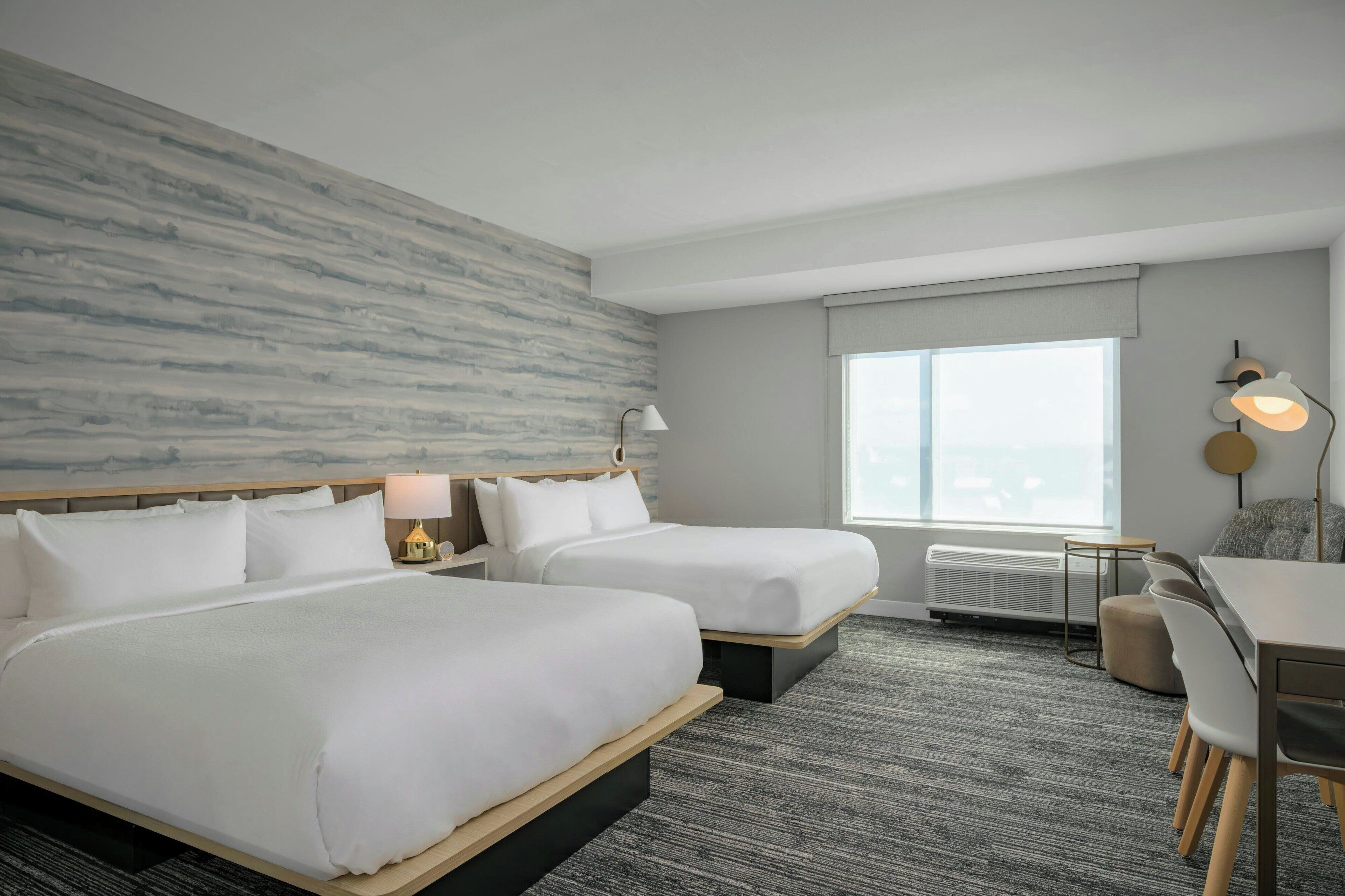 A hotel room with two beds, a desk, a chair, a lamp, and a large window.