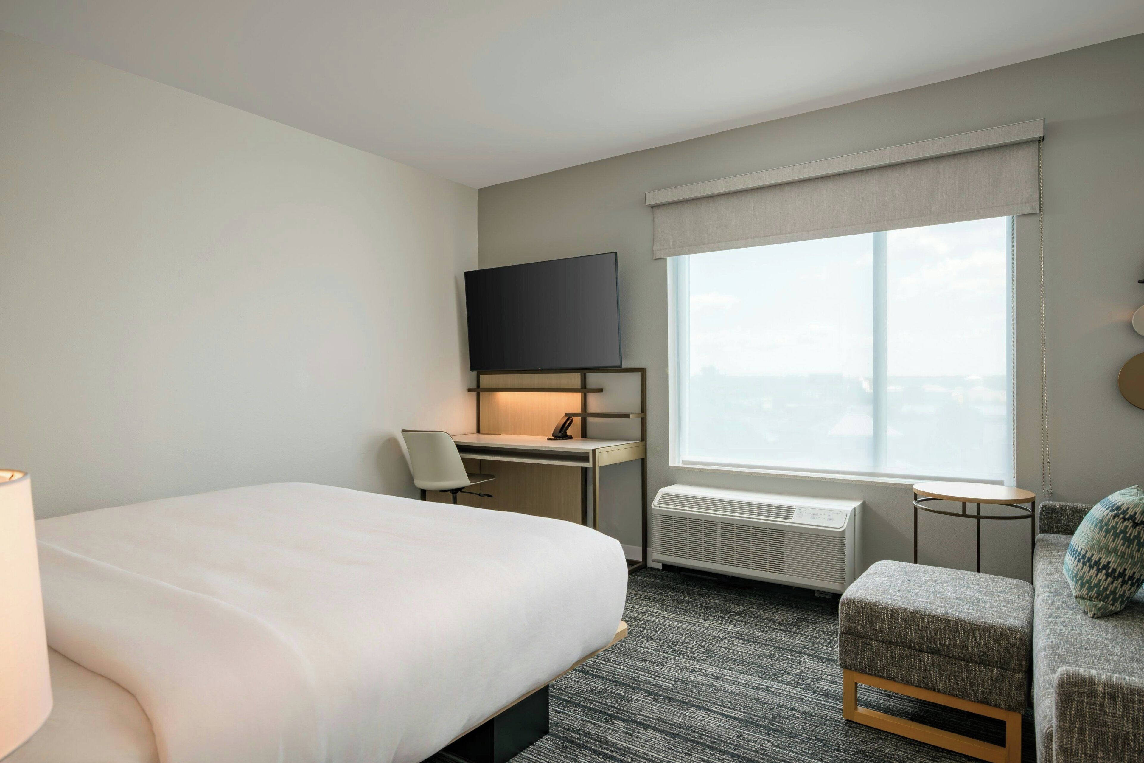 A hotel room with a bed, a desk with a chair, a television, a window with a view, and a heater.