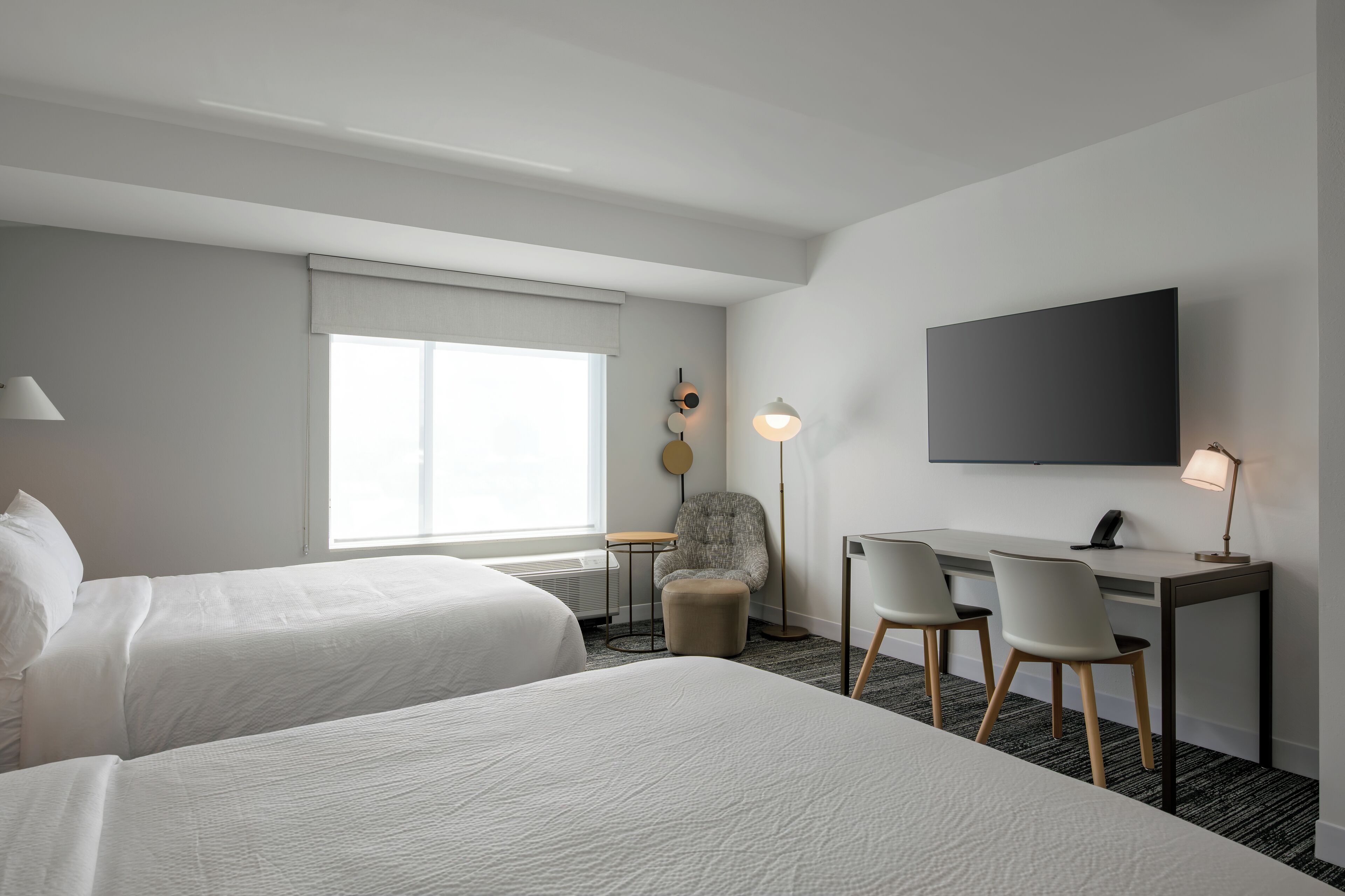 A hotel room with two beds, a desk with chairs, a flat-screen TV, a lamp, and a window with a view.