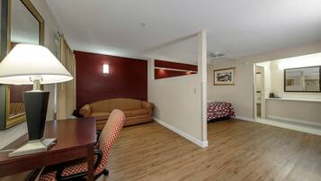 Una habitación con suelo de madera, un escritorio con lámpara, una silla a cuadros, un sofá rojo, una cama con colcha roja y blanca a cuadros, y un espejo en la pared.