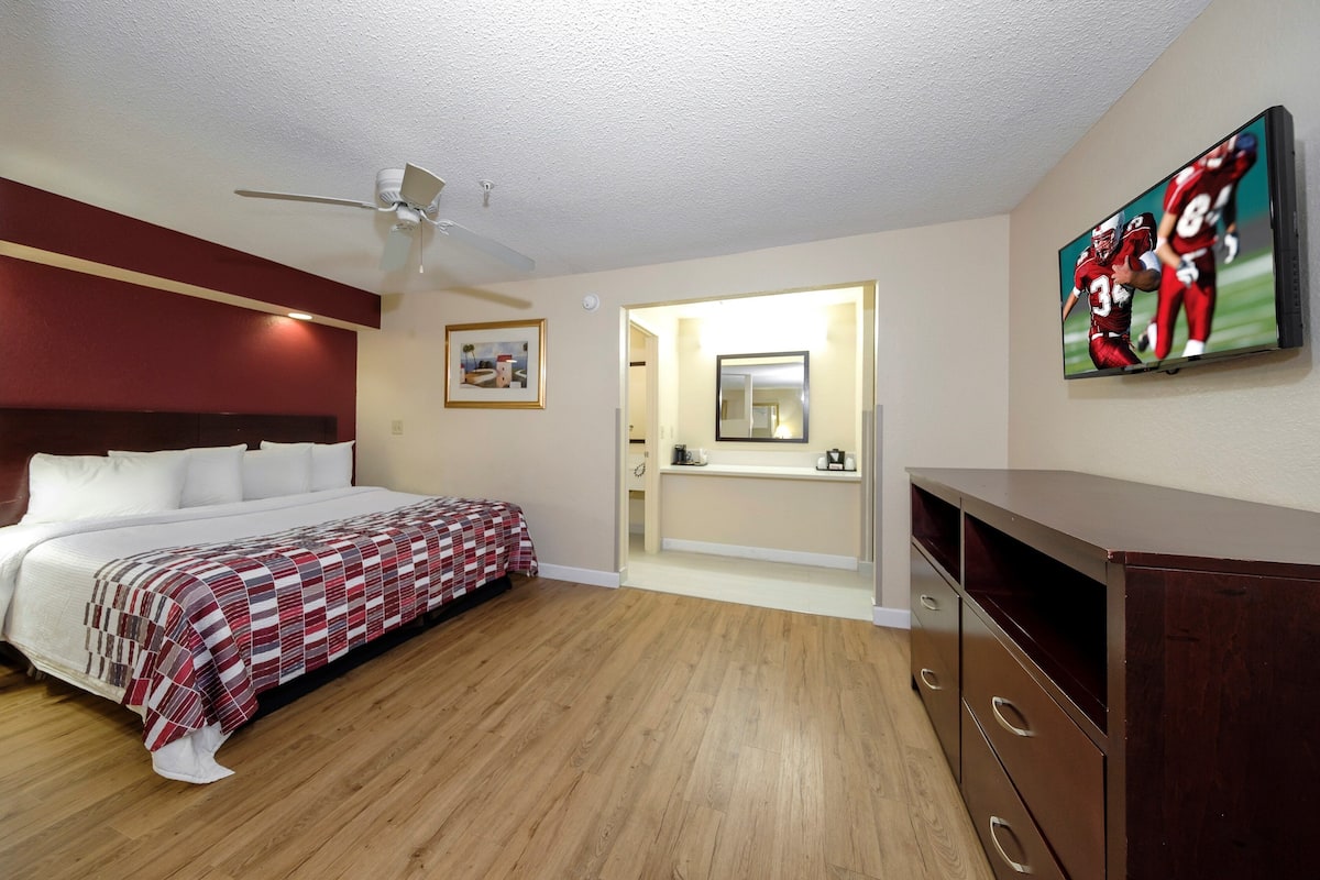Ein Schlafzimmer mit Bett, Deckenventilator, Fernseher, Kommode und einem durchgegangenen Tür erkennbaren Badezimmer.