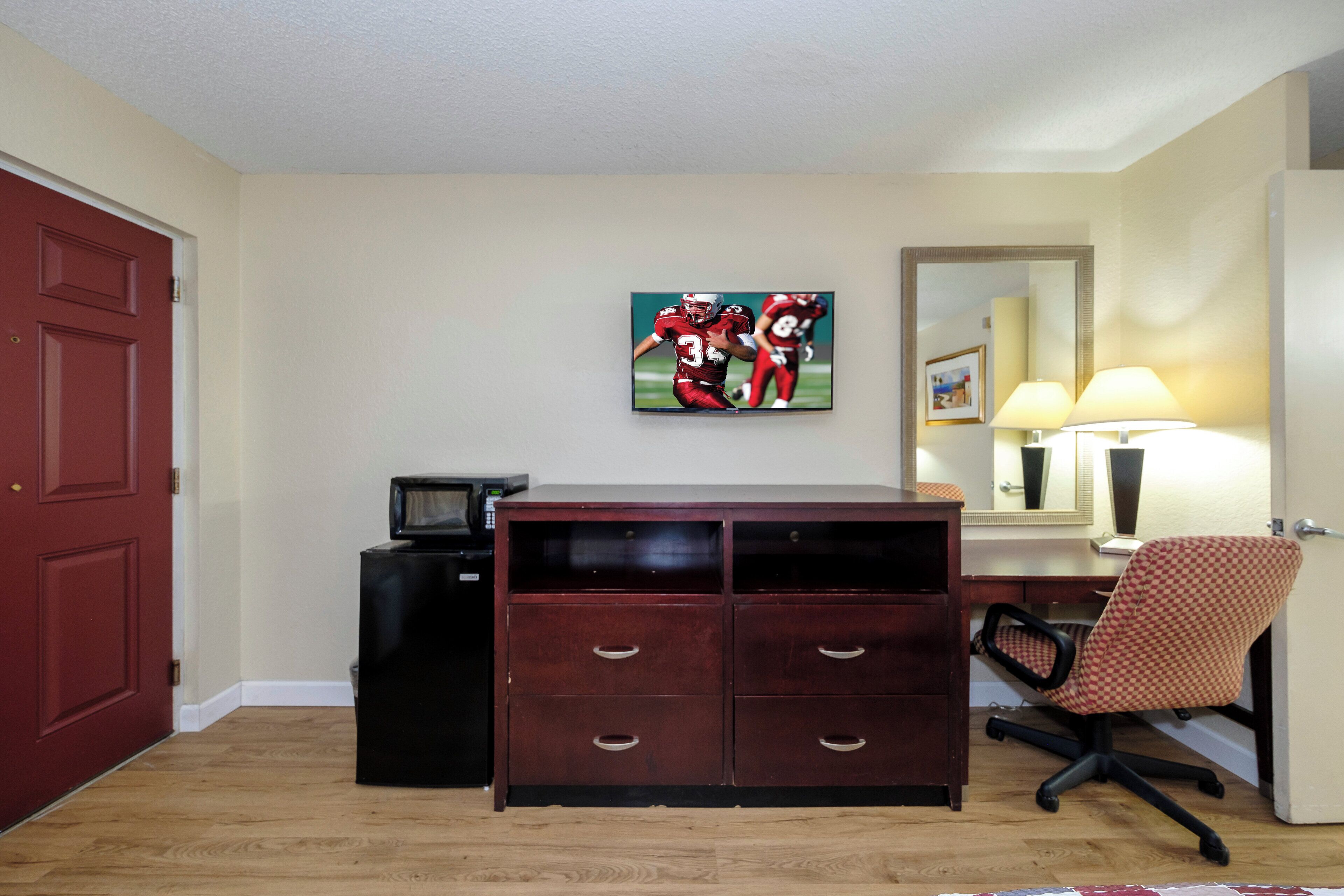 A hotel room with a wooden desk, a chair, a television mounted on the wall, and a red door.