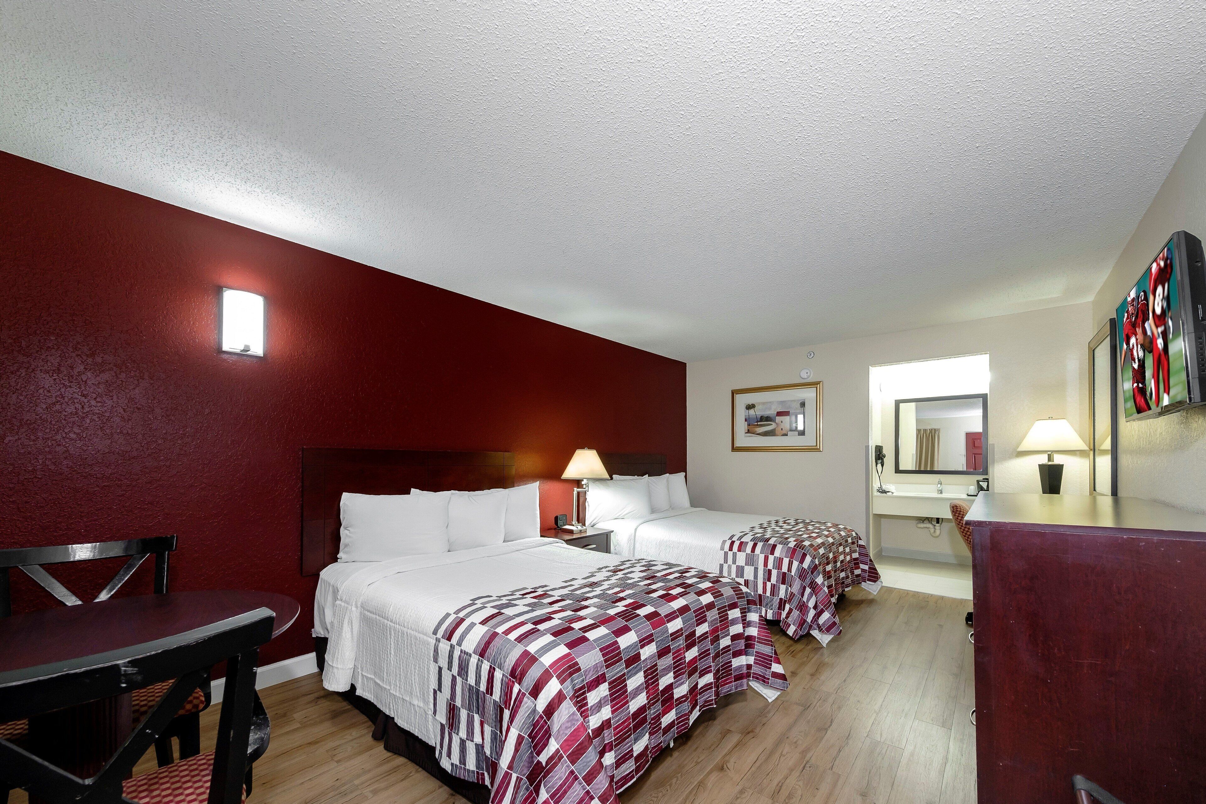 A hotel room with two beds, a desk, a chair, a small table, a lamp, and a mirror.