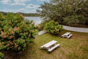 A grassy backyard with a white picket fence, two picnic tables, and dense foliage.