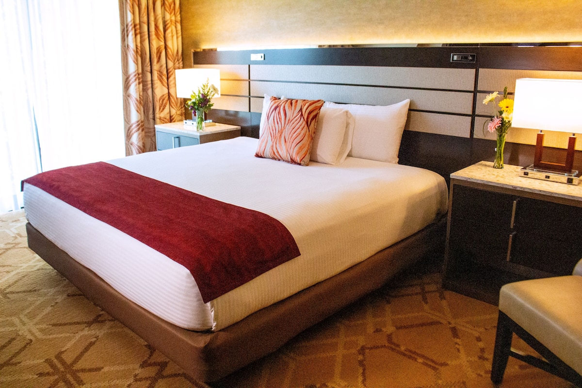 A hotel room with a large bed, a nightstand with a lamp, a chair, and a wall-mounted TV.