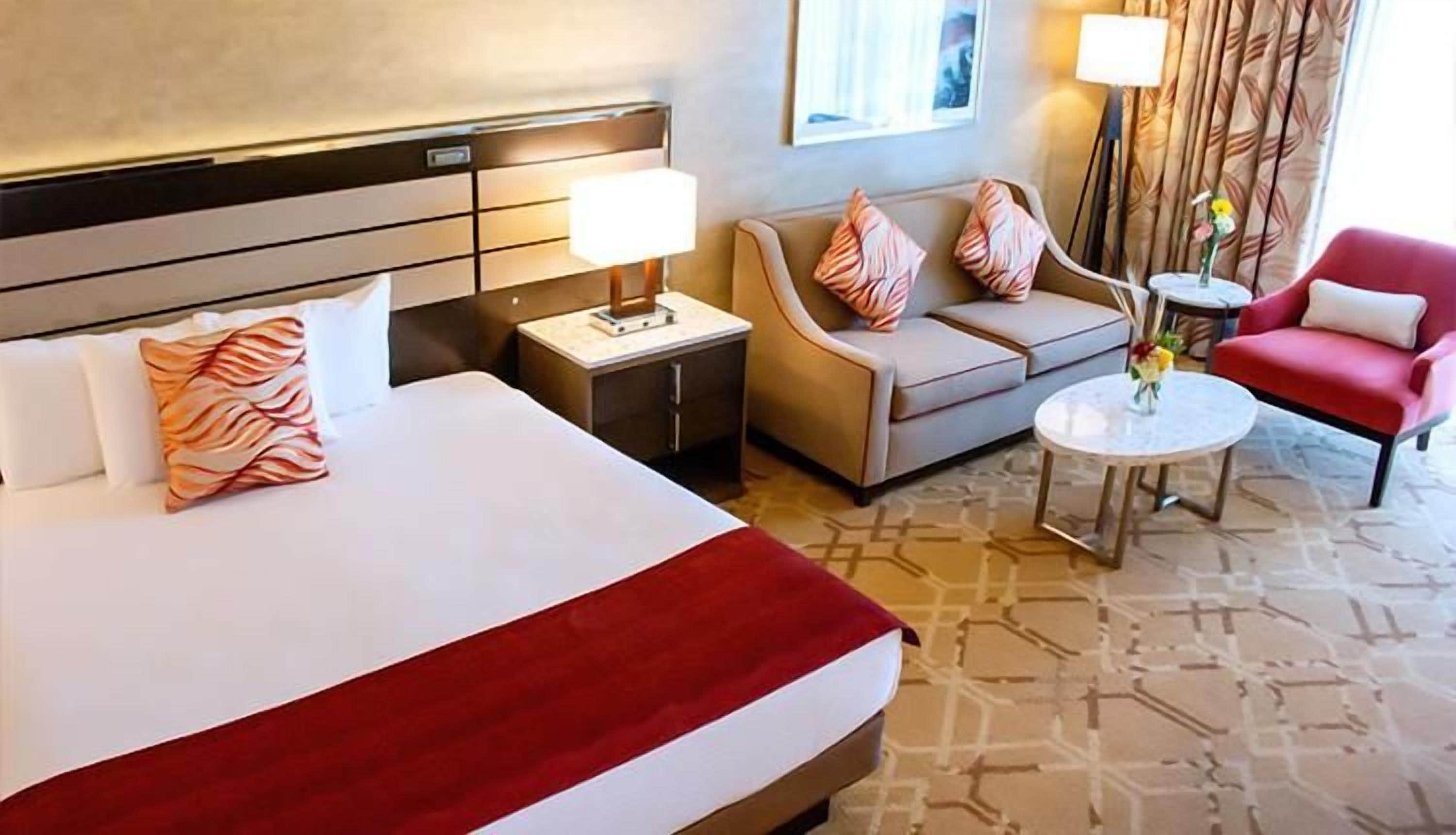 Una habitación de hotel con cama, sofá, sillón rojo, mesita y lámpara.