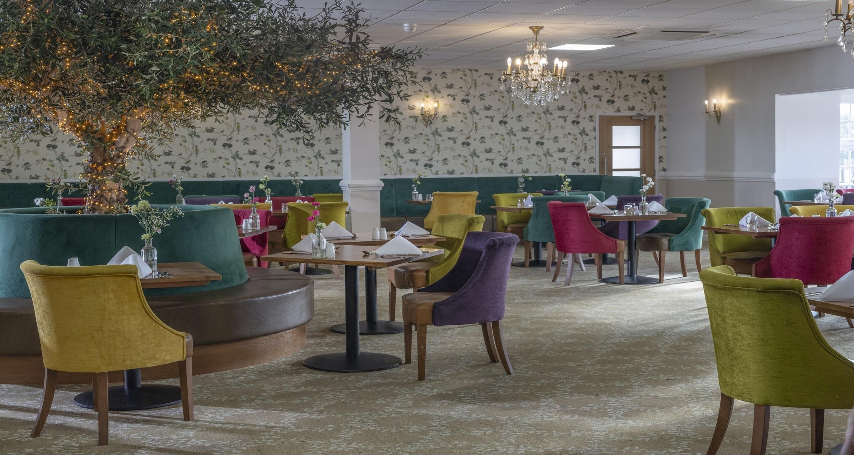 Un restaurant avec des tables rondes et des chaises colorées, un grand arbre illuminé et un papier peint à motifs floraux.