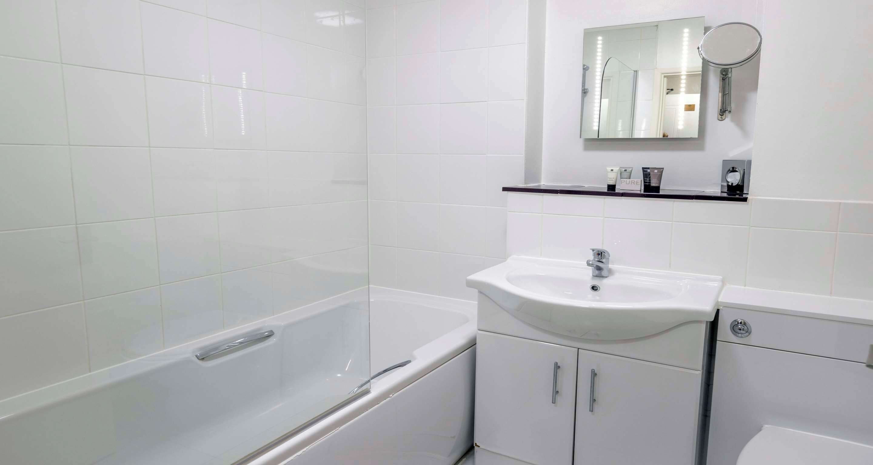 A bathroom with white tiles, a bathtub, a sink, a mirror, and a shelf with toiletries.