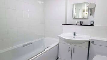 A bathroom with white tiles, a bathtub, a sink, a mirror, and a shelf with toiletries.