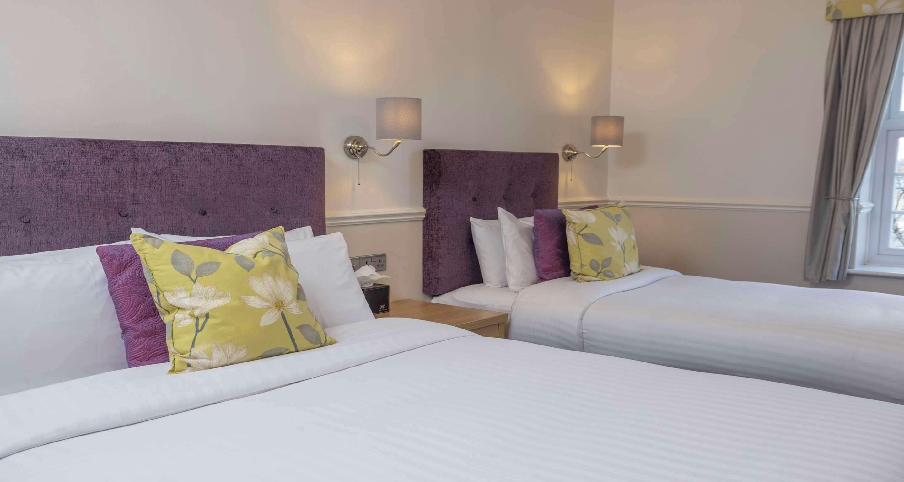 Habitación de hotel con dos camas, cada una con ropa blanca y cabeceros morados. Hay dos lámparas de pared a cada lado de las camas.