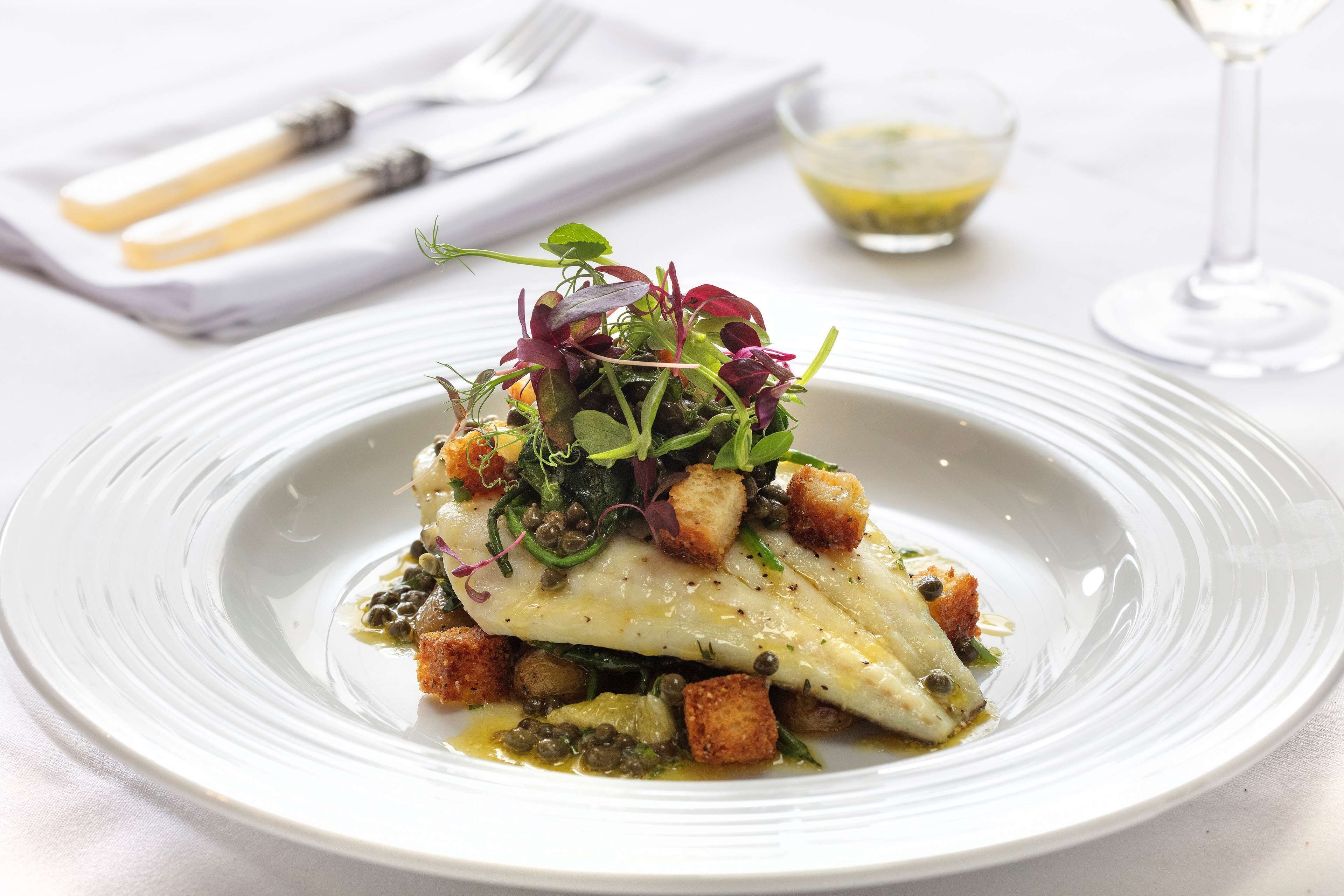 A white plate with a fish dish, garnished with greens and capers, accompanied by croutons and a small bowl of sauce.