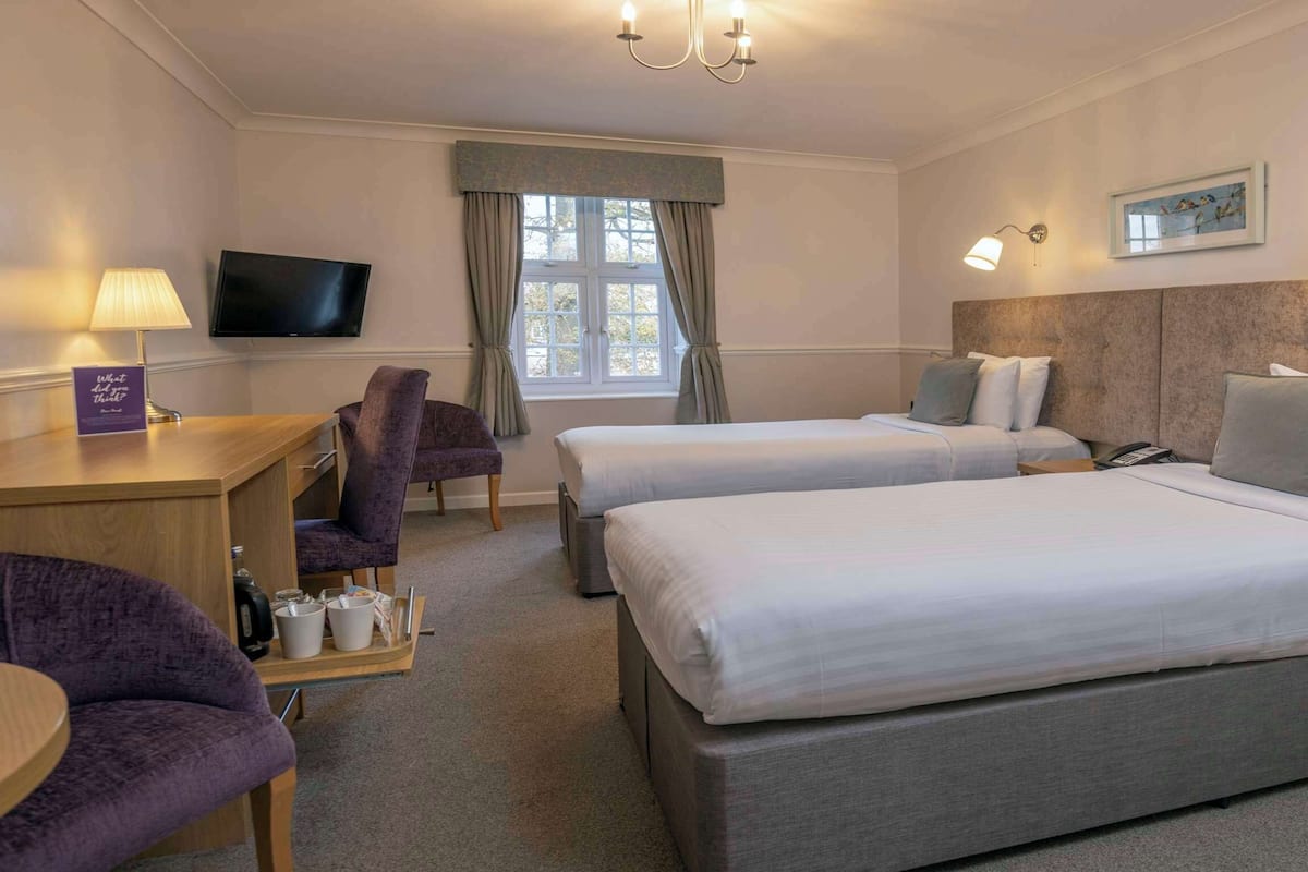A hotel room with two beds, a desk with a lamp, a chair, a TV, and a window with curtains.