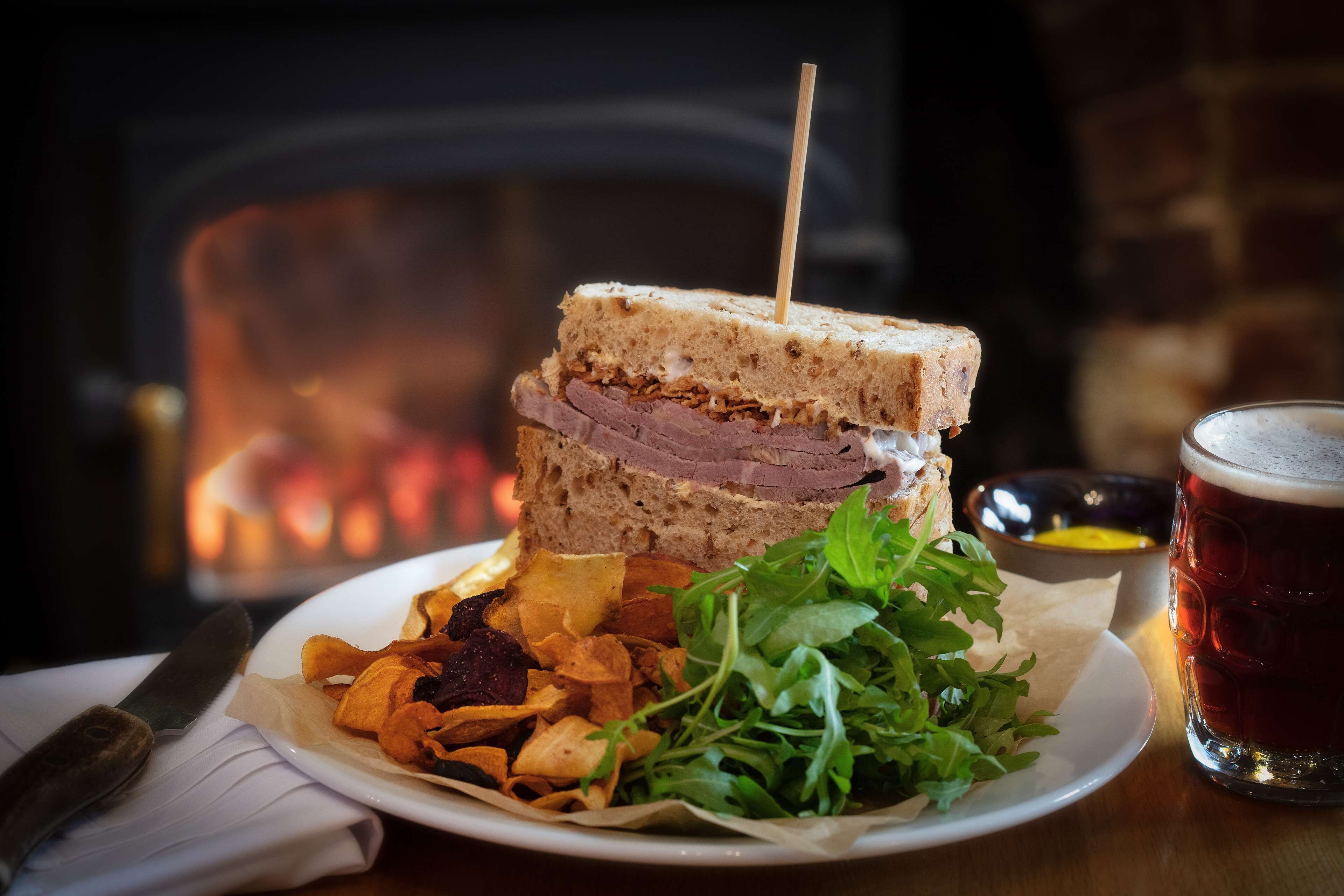 A sandwich with a toothpick, a plate of chips, a glass of beer, and a napkin with a knife on a wooden table.