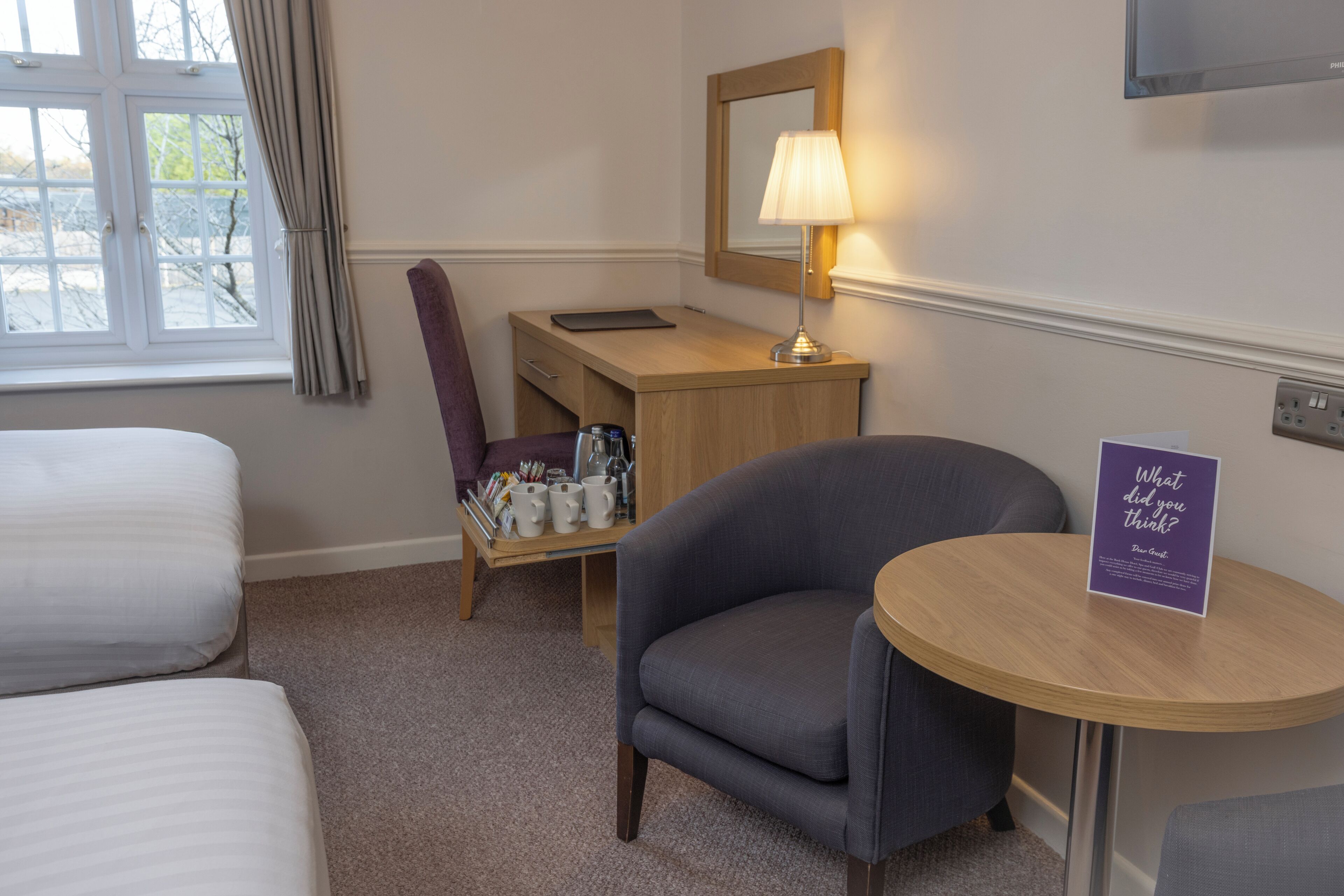 A hotel room with a bed, a desk with a lamp, a chair, and a small table with a sign.