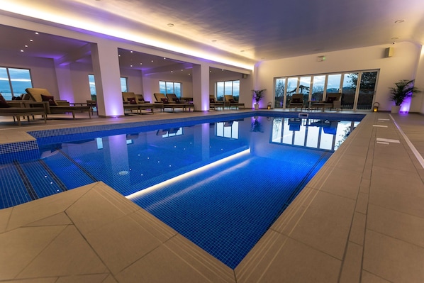 An indoor swimming pool with clear blue water, surrounded by lounge chairs and large windows offering views of the outdoors.