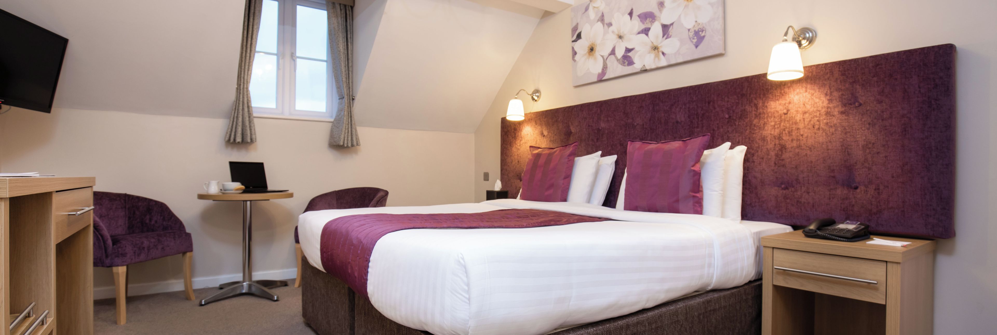 A hotel room with a bed, a desk, a chair, a small table, a lamp, and a painting on the wall.