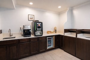 Una cocina moderna con gabinetes de madera oscura, un lavamanos de acero inoxidable, una cafetera, un dispensador de bebidas y un refrigerador.
