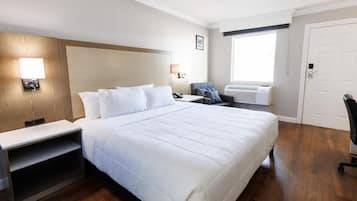 A hotel room with a large bed, bedside tables, a desk, a chair, an air conditioning unit, and a door.