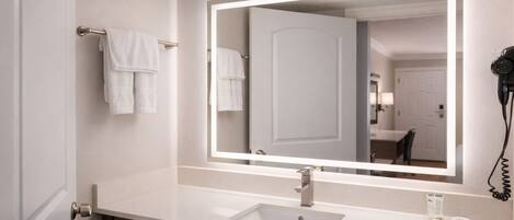 A modern bathroom with a white sink, a large mirror with integrated lighting, and a hairdryer on the counter.
