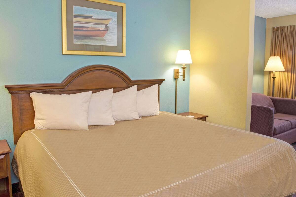 Una habitación de hotel con una cama grande, una mesita de noche, una lámpara, una silla y un cuadro en la pared.