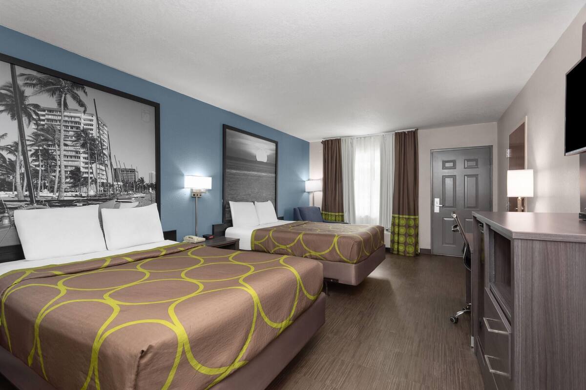 Habitación de hotel con dos camas, un escritorio, un televisor y un ventanal con cortinas.