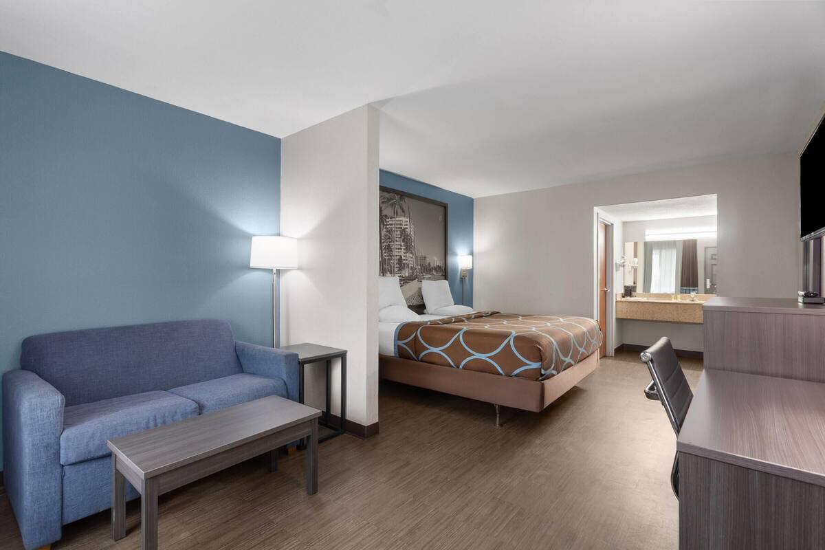 Habitación de hotel con cama, sofá, escritorio, lámpara y baño visible a través de una puerta abierta.