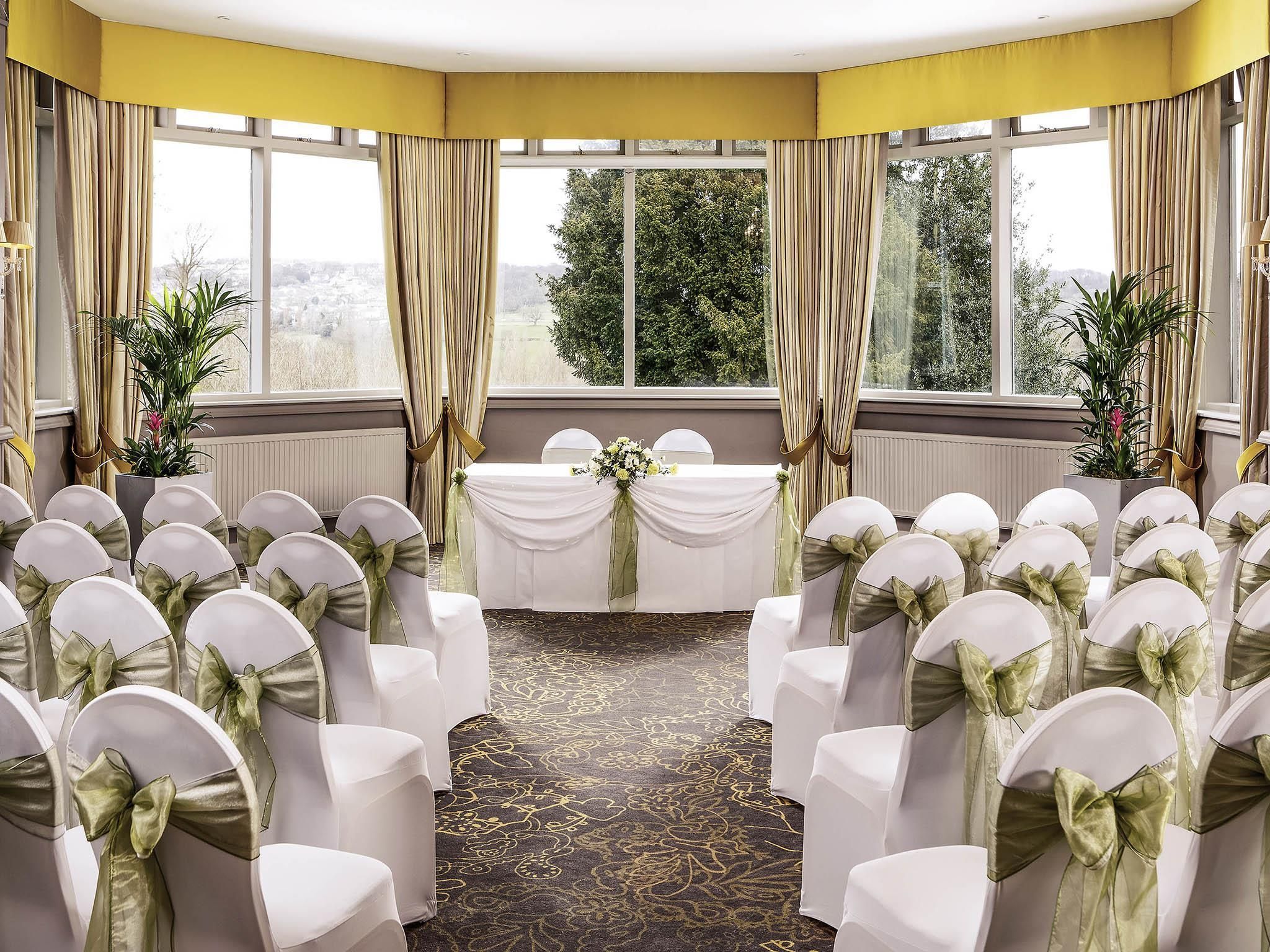 A conference room with rows of white chairs, a table with a white tablecloth, and large windows with curtains.