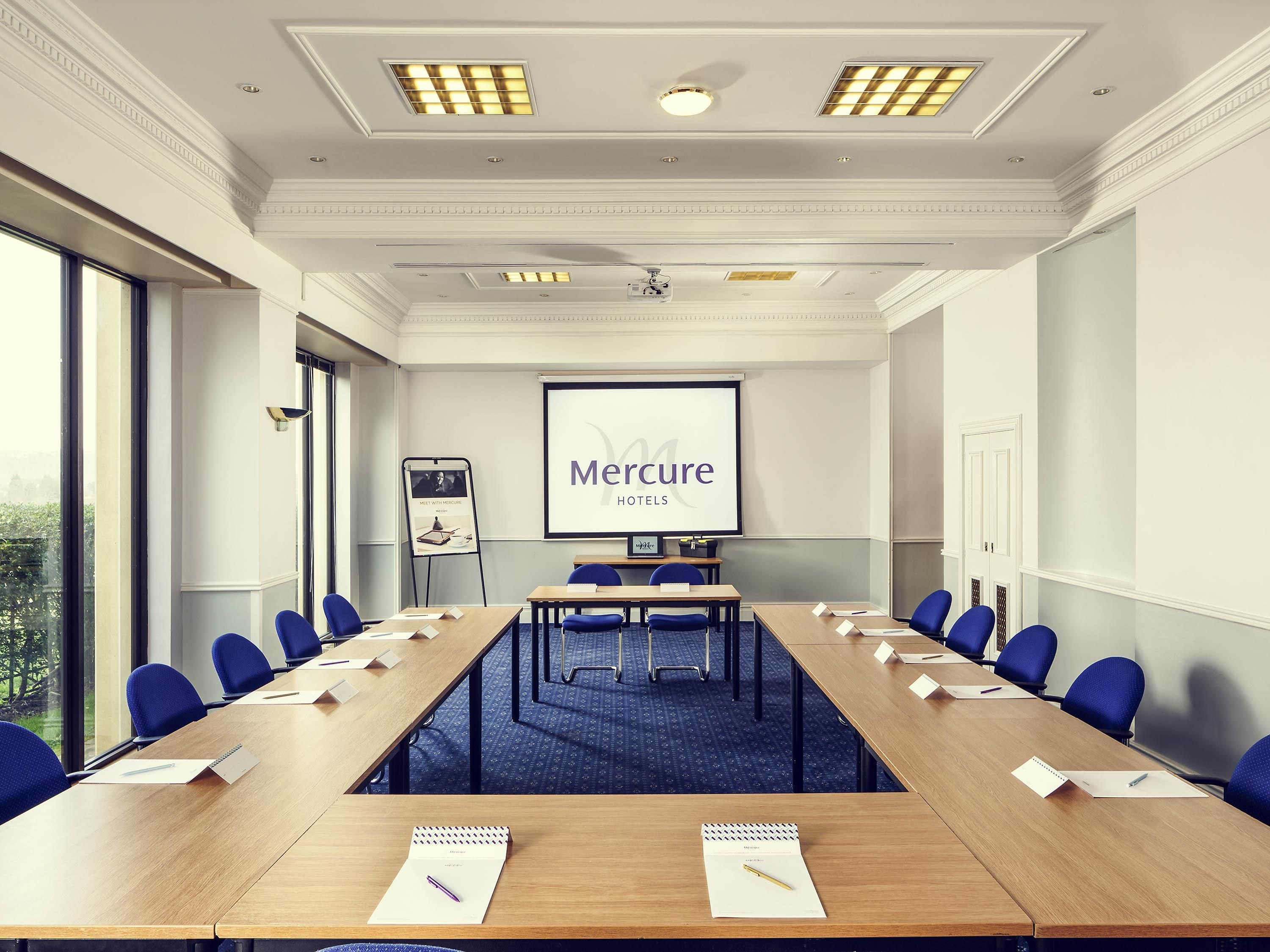A conference room with a long table, blue chairs, a projector screen displaying 'Mercure Hotels,' and a window with a view.