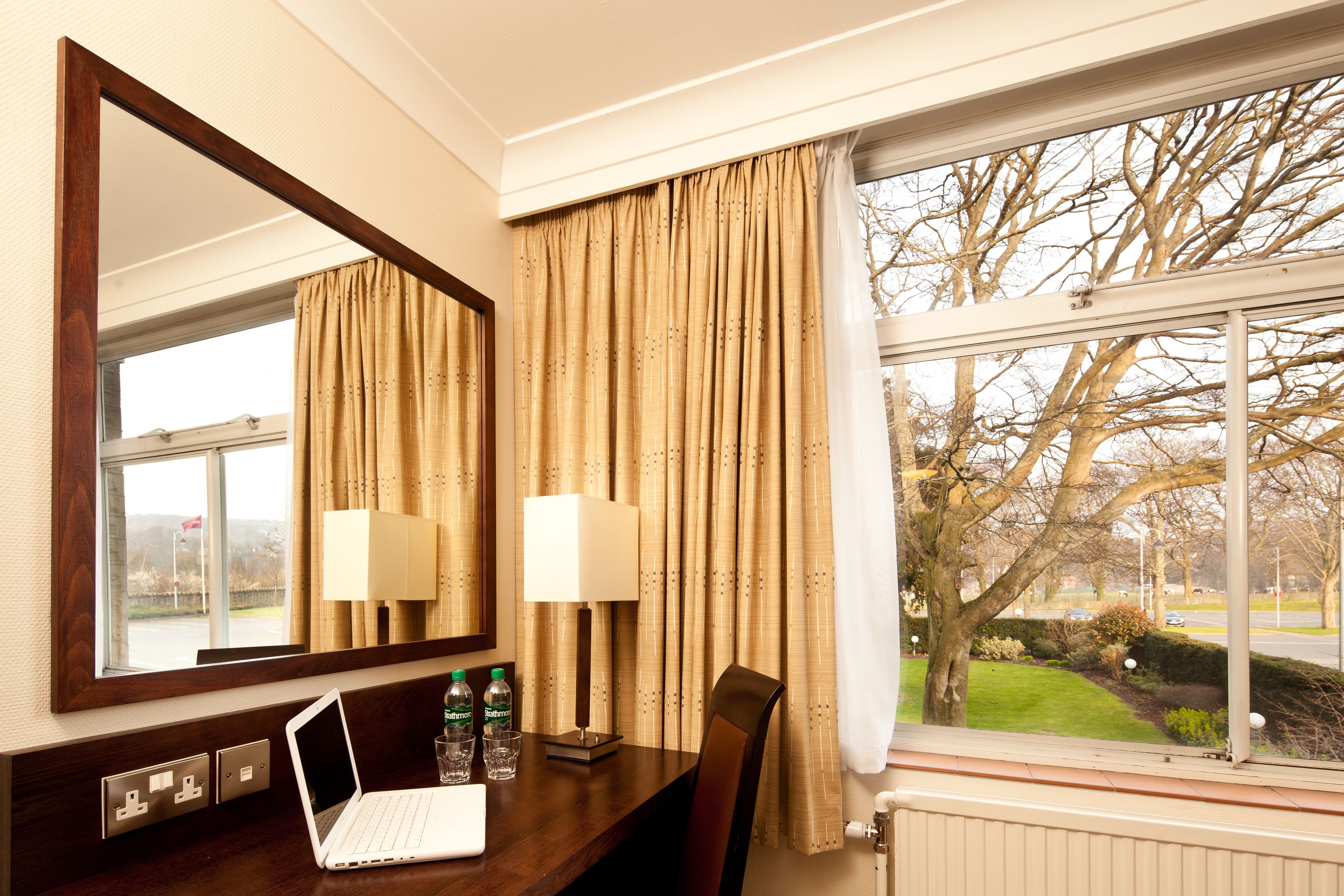 Habitación de hotel con un escritorio de madera, una computadora portátil, dos botellas de agua, una lámpara y vistas a una calle arbolada a través de una ventana con cortinas.