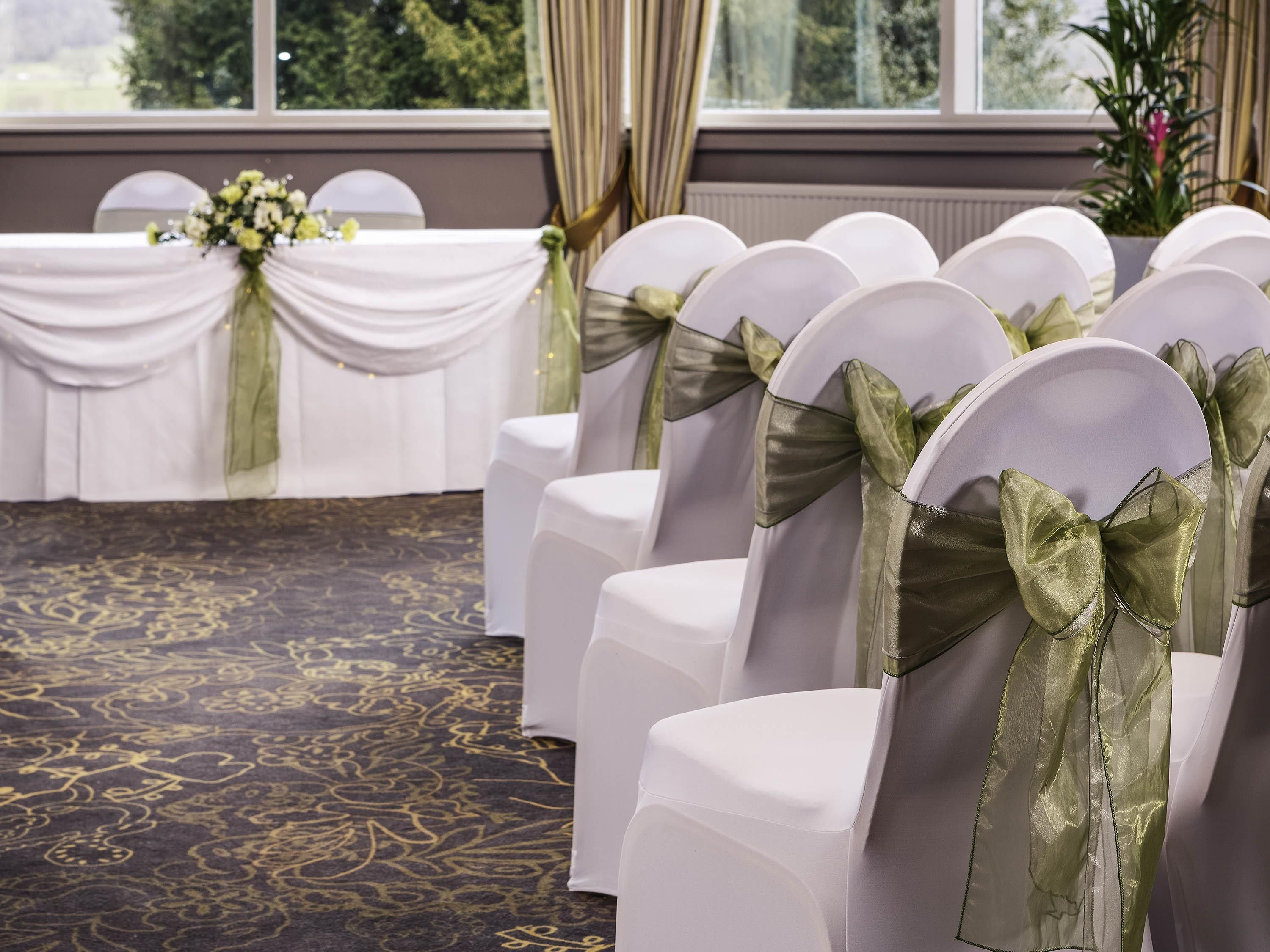 Rows of white chairs with green sashes, a table with a white tablecloth and floral centerpiece, and a patterned carpet.